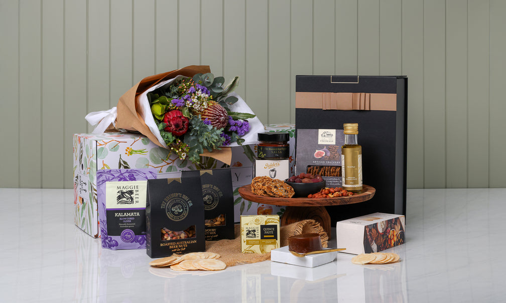 Gourmet gift basket with flowers, snacks, and a bottle on a white surface.