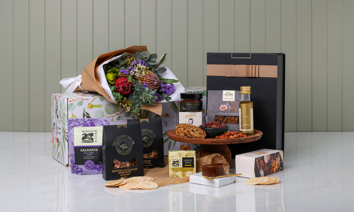 Gourmet gift basket with flowers, snacks, and a bottle on a white surface.