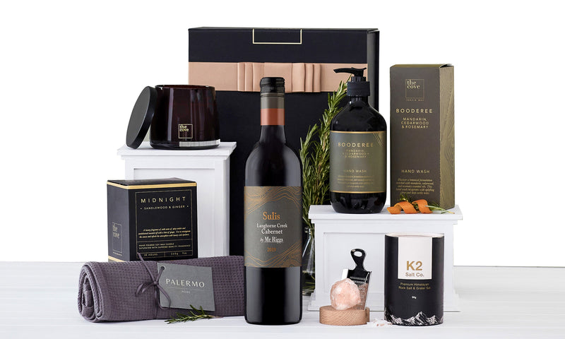 A gift hamper set including a bottle of red wine, a box of chocolates, a wine glass, and other assorted gourmet food items.